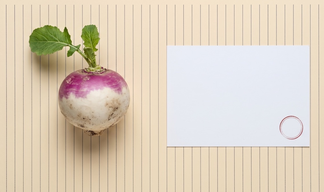 Um nabo verdadeiro, com terra e folhas, ao lado de um postal em branco — registo fotográfico.